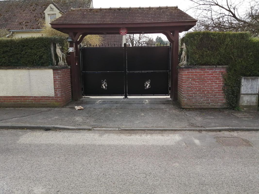 Portail en métal noir sous un abri de voiture en bois, à côté d'un mur de briques, donnant sur une allée et une route.
