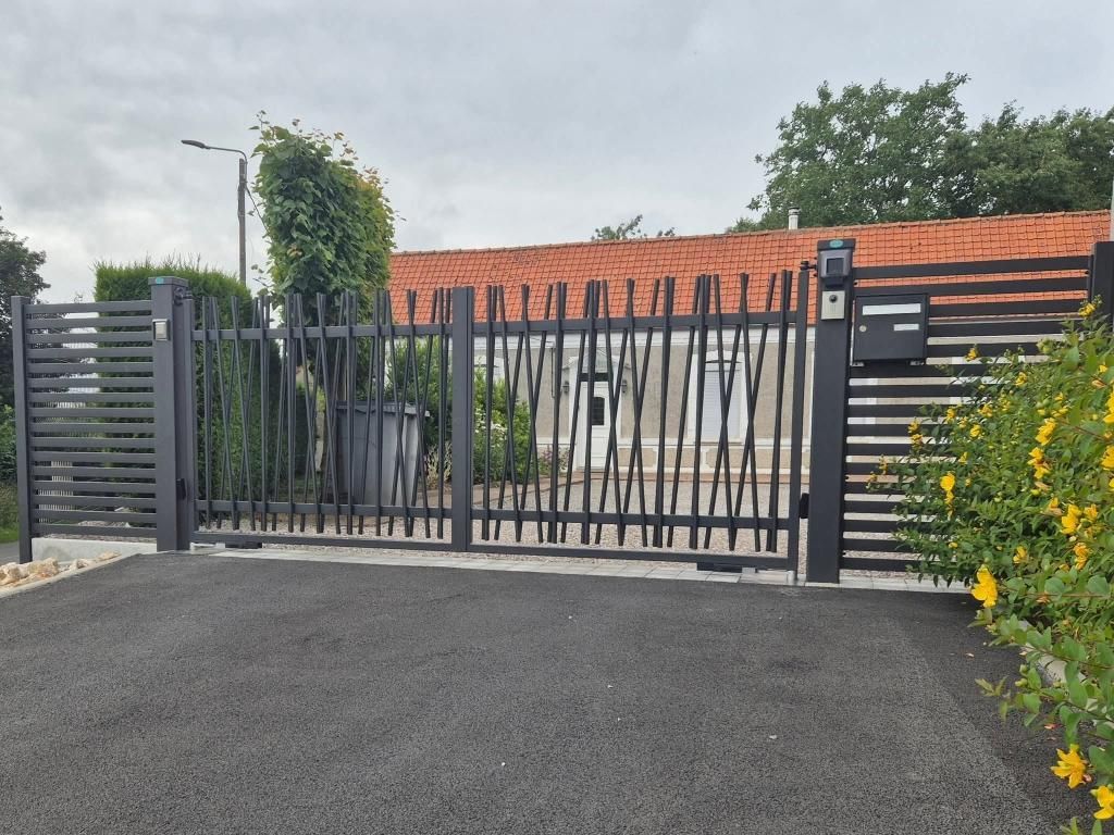 Un portail en fer forgé noir se dresse devant une maison en briques au toit de tuiles et à la terrasse pavée, donnant sur une cour gazonnée.