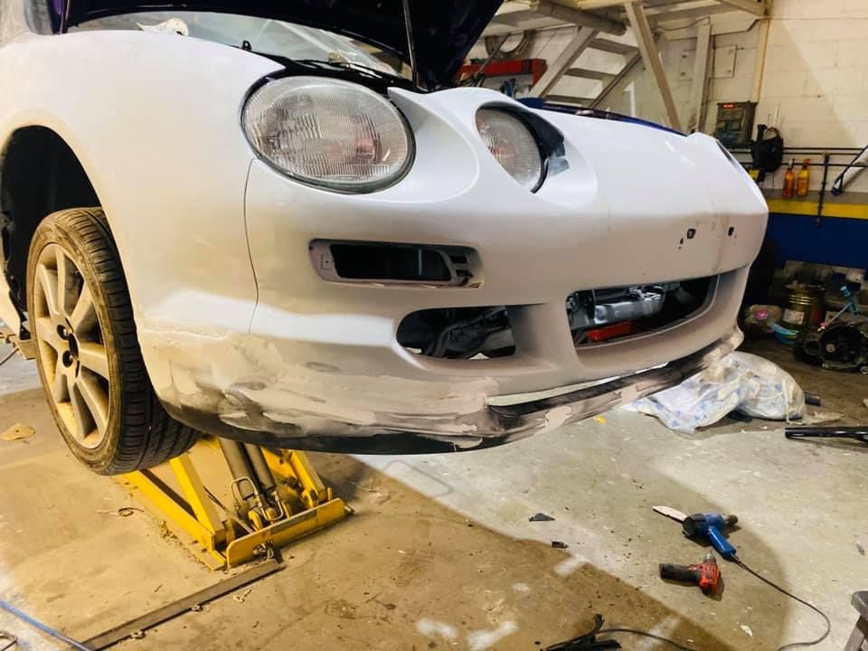 El parachoques delantero de un coche blanco sobre un elevador, en reparación en un garaje.