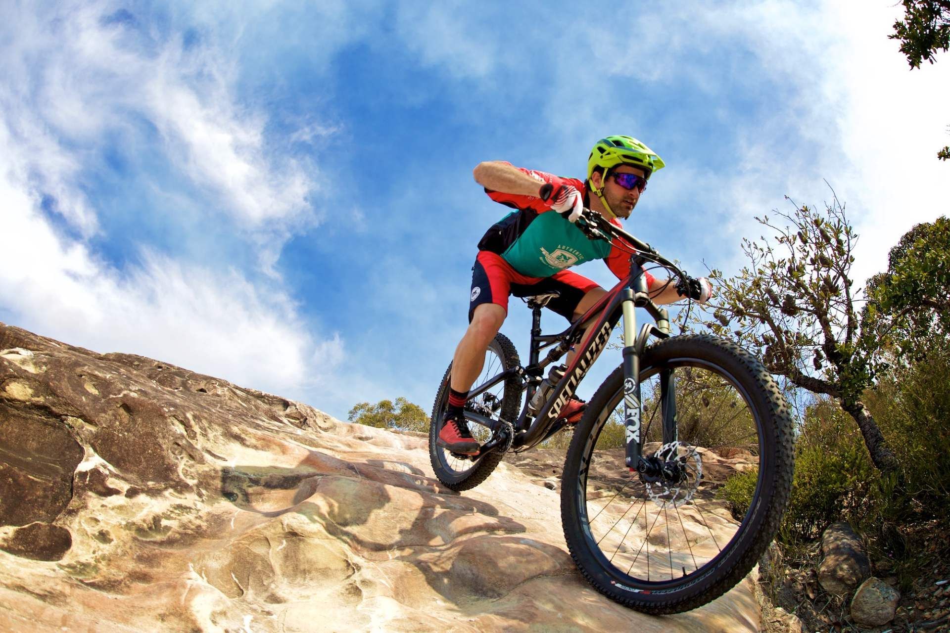 Mountainbiker mit neonfarbenem Helm fährt unter blauem Himmel einen felsigen Pfad hinunter