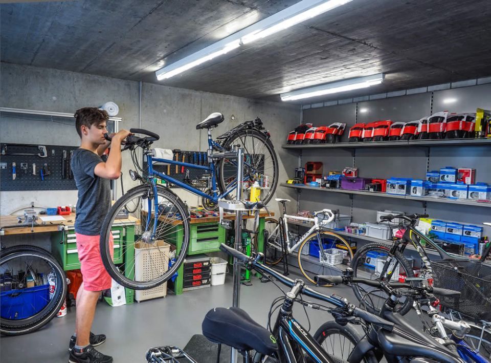 Eine Person repariert ein Fahrrad in einer vollgestellten Fahrradwerkstatt mit Werkzeugen, Ersatzteilen und mehreren Fahrrädern in Regalen.