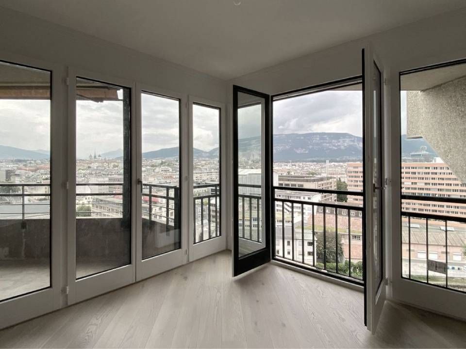 Intérieur d'un appartement moderne vide, avec de grandes fenêtres et un balcon donnant sur la ville et les montagnes.