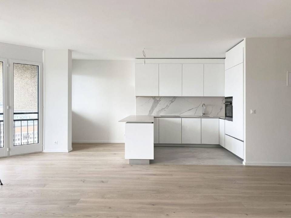 Cuisine et séjour modernes et blancs, vides, avec parquet clair et porte-fenêtre coulissante.