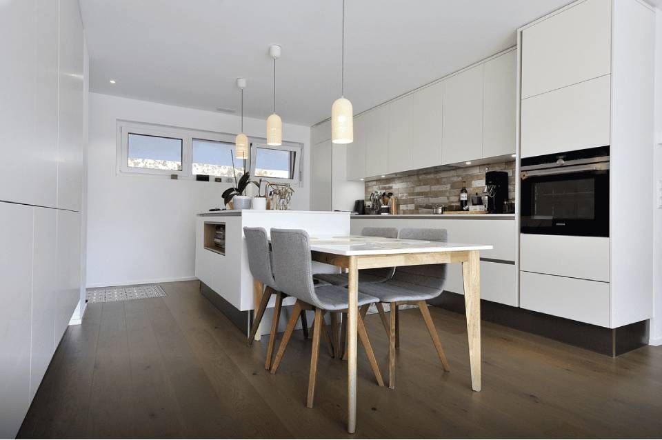Cuisine et salle à manger modernes blanches avec suspensions lumineuses, îlot central et table en bois sur sol foncé
