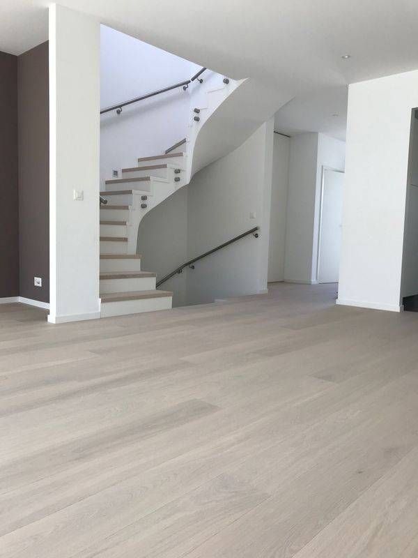 Salon moderne vide avec parquet clair, murs blancs et escalier à rampe en verre.