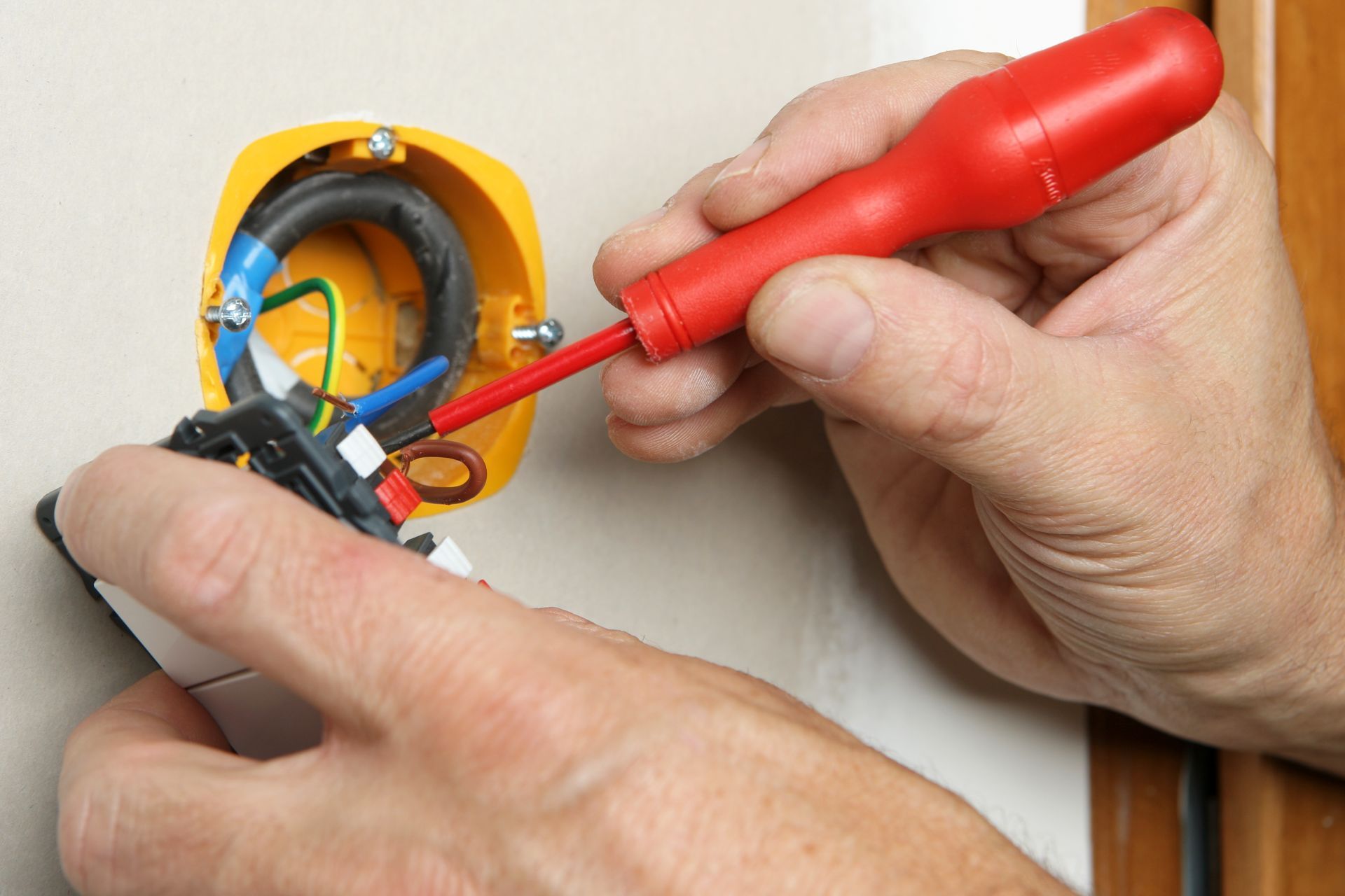 Des mains utilisent un tournevis rouge pour réparer le câblage électrique d'une prise murale.