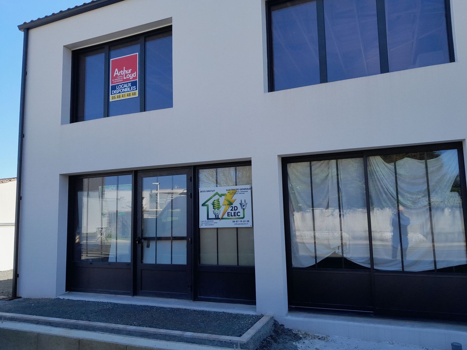 Bâtiment moderne blanc de deux étages avec fenêtres sombres et portes vitrées en façade.