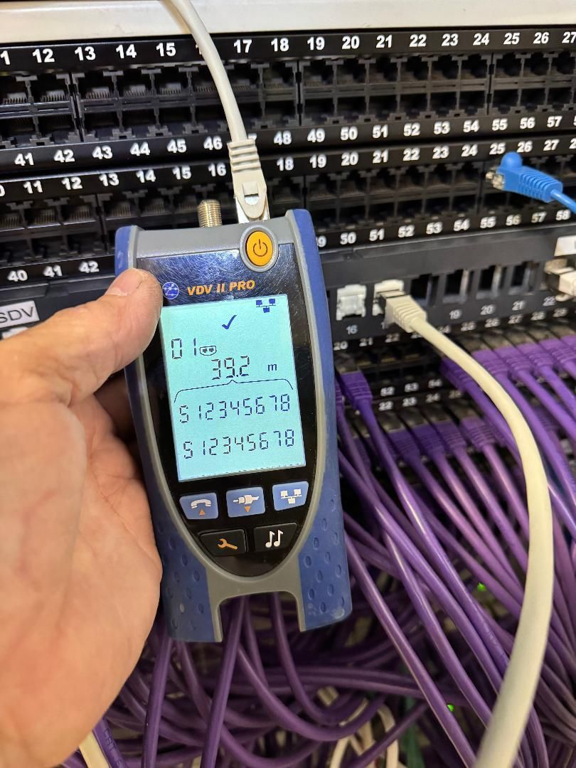 Main tenant un testeur de câbles réseau devant un panneau de brassage avec des câbles et des ports violets.