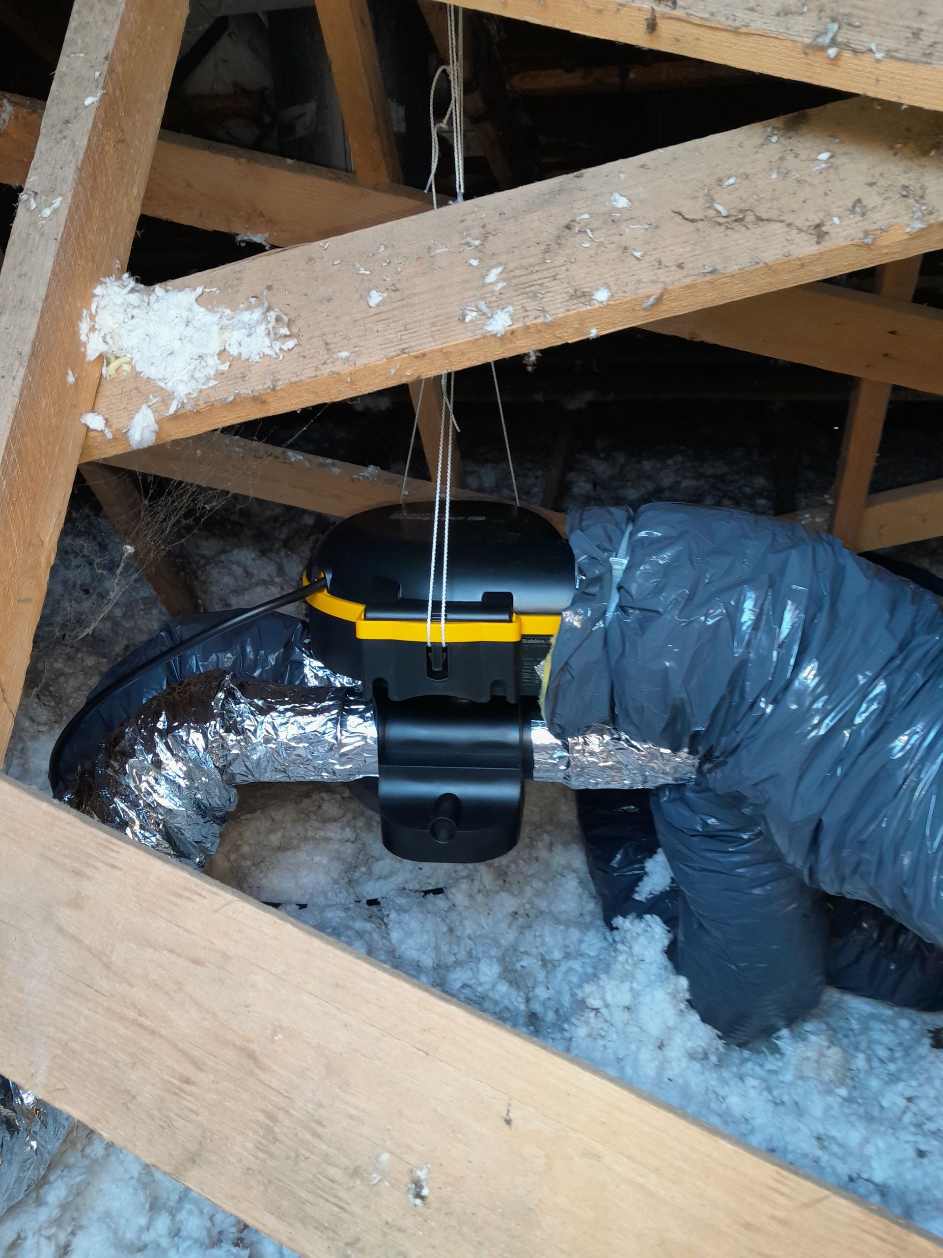 Grenier avec conduits isolés et un appareil noir monté sur un conduit argenté, entouré de poutres en bois.