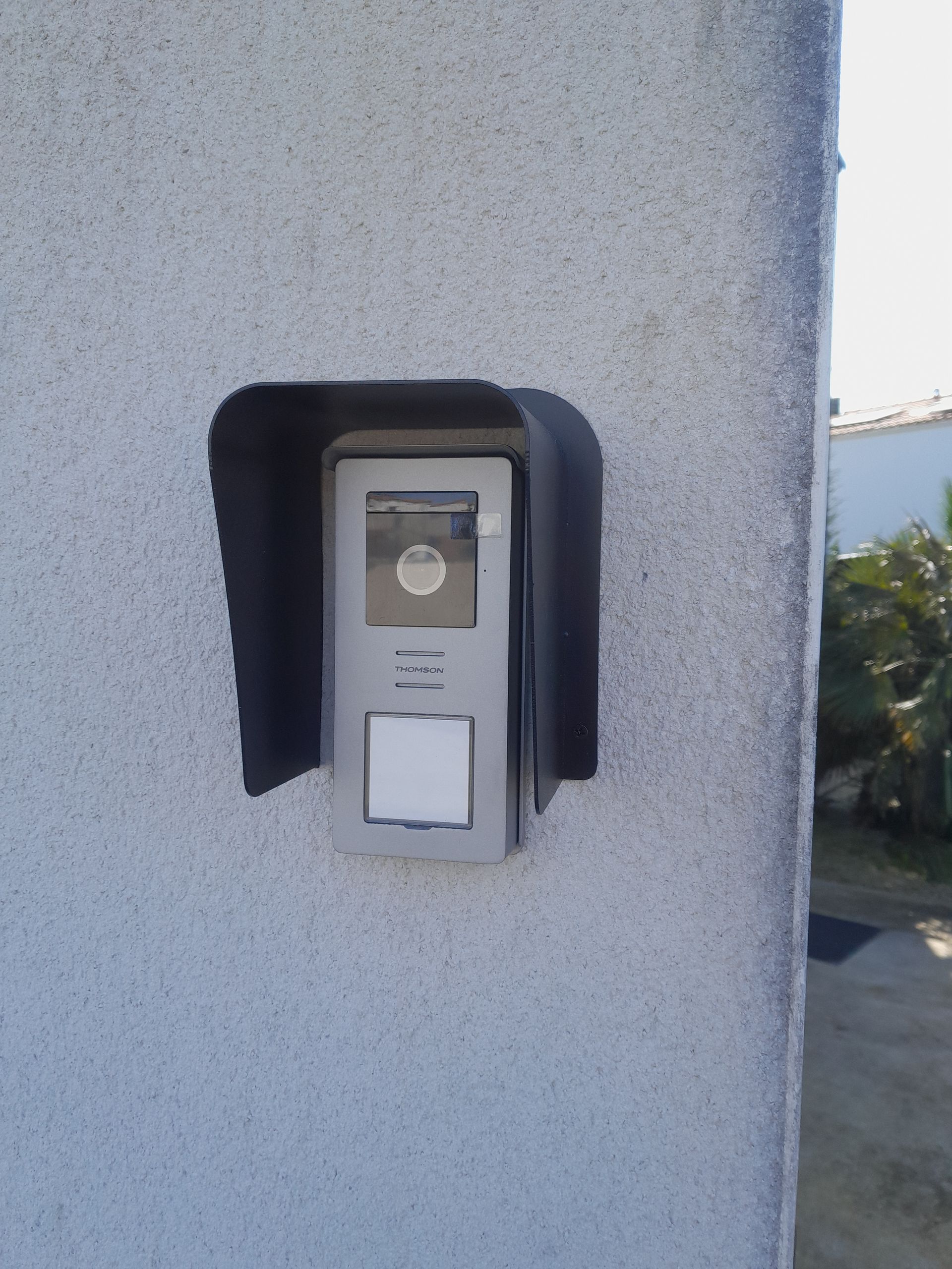 Sonnette d'interphone murale fixée sur un mur extérieur texturé, avec une petite caméra et un bouton.