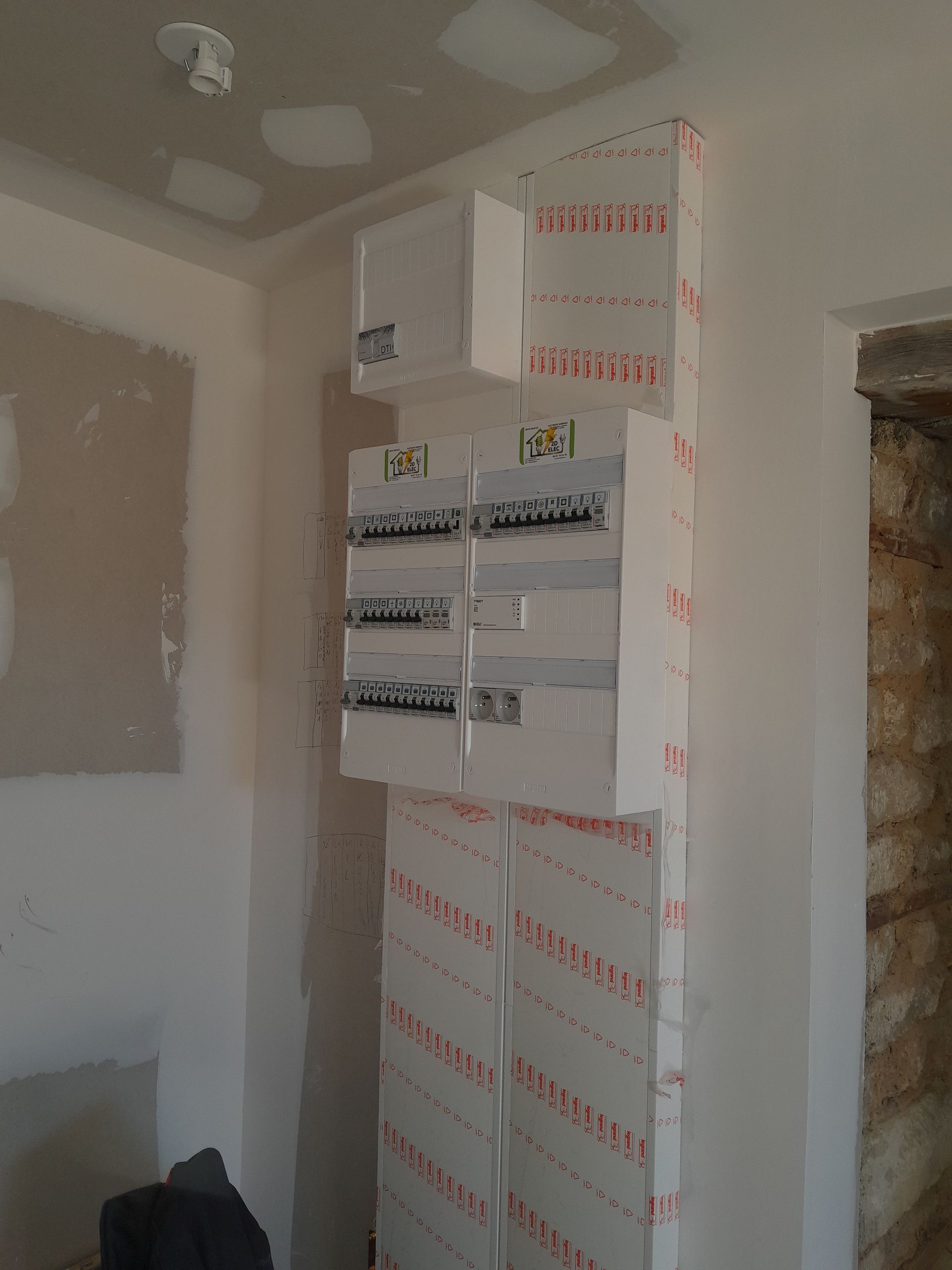 Tableau électrique et boîtes de disjoncteurs fixés sur un mur en plaques de plâtre non finies dans une pièce en rénovation.