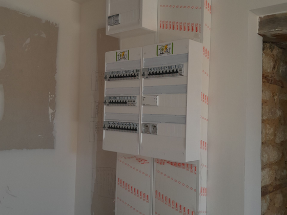 Tableau électrique et boîtes de disjoncteurs fixés sur un mur en plaques de plâtre non finies dans une pièce en rénovation.