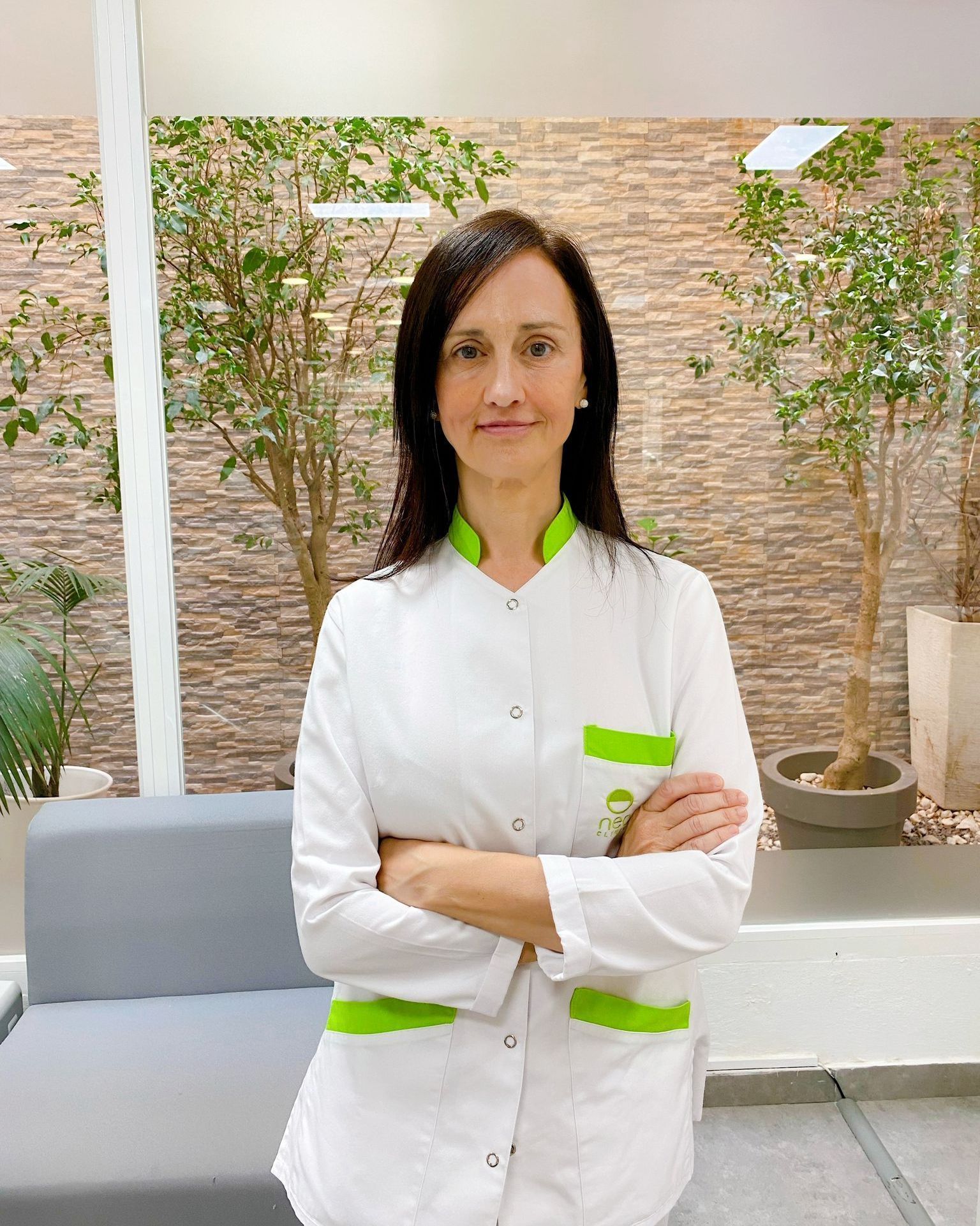 Una mujer con una bata de laboratorio blanca está parada con los brazos cruzados frente a un sofá.