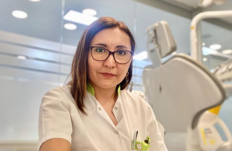 Una mujer que lleva gafas y una bata blanca está sentada frente a una silla dental.