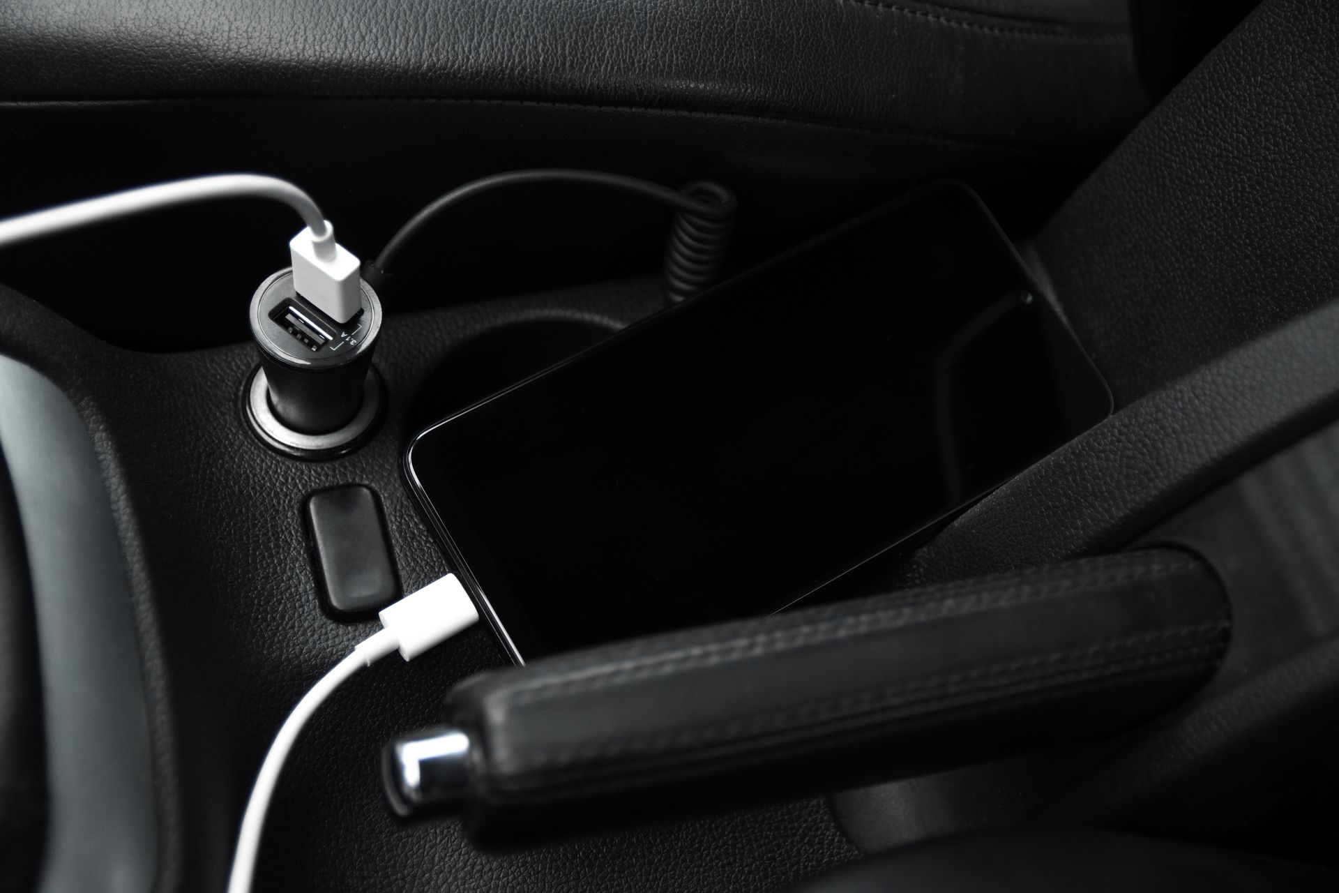 Téléphone portable noir en charge dans la voiture, connecté à un chargeur USB placé dans un porte-gobelet, près d'un frein à main noir.