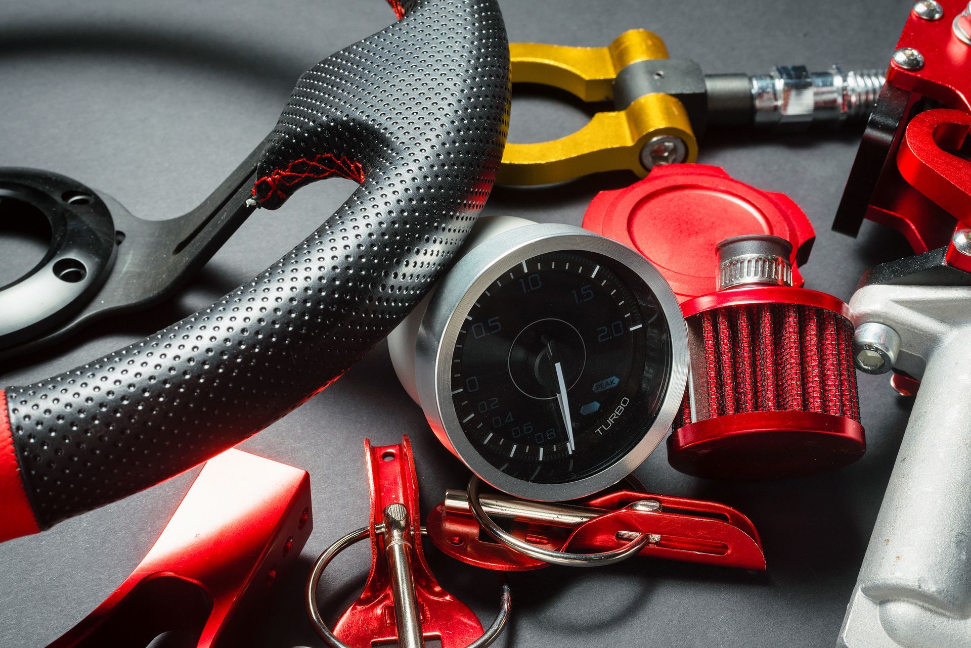 Gros plan sur des pièces automobiles : volant noir, cadrans, accessoires rouges et dorés, fond gris.