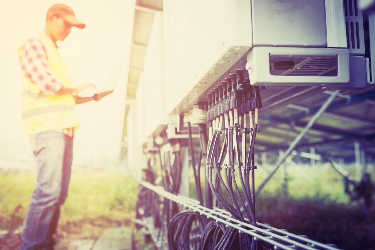 Un técnico con chaleco de seguridad inspecciona el cableado de una instalación de paneles solares al aire libre. Los cables salen de una gran caja metálica.