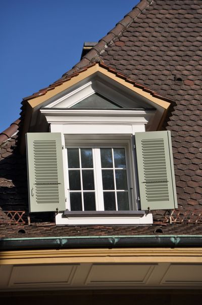 Lucarne aux volets verts sur un toit en tuiles brunes.