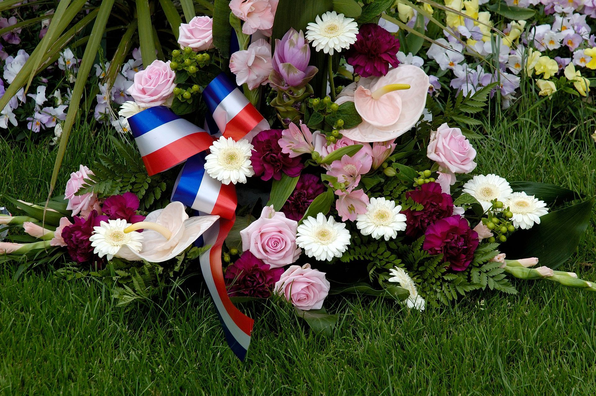 Couronne de fleurs roses, blanches et jaunes, ornée d’un ruban tricolore, posée sur de l’herbe verte