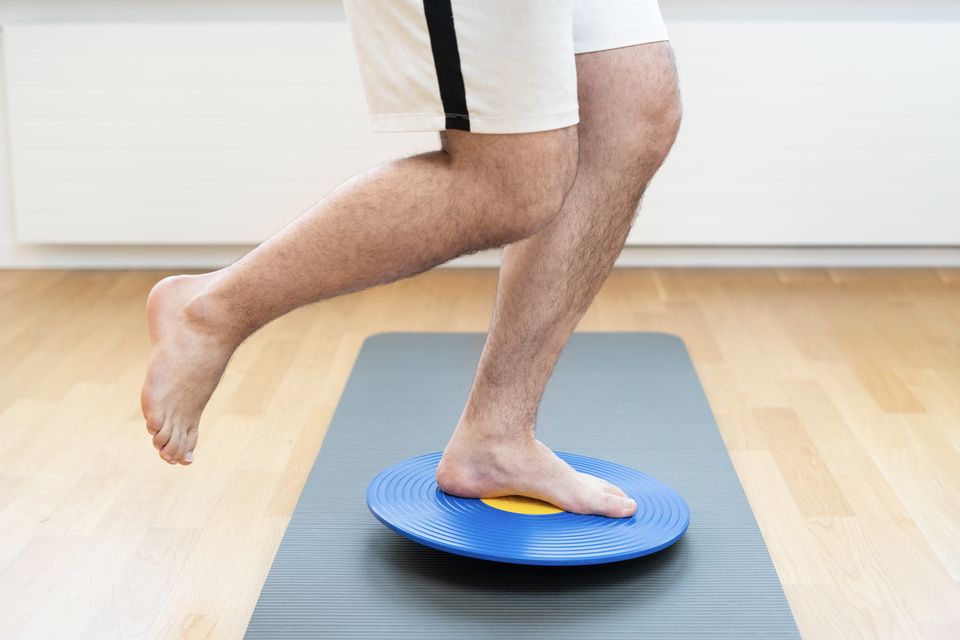 Ein Mann steht auf einem Balance Board auf einer Yogamatte. Foto von der Physiotherapie Kriens