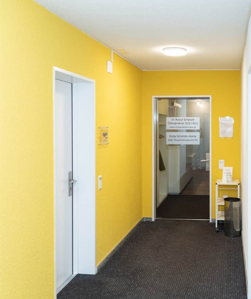 Ein Flur mit gelben Wänden und schwarzem Teppich. Foto von der Physiotherapie Kriens