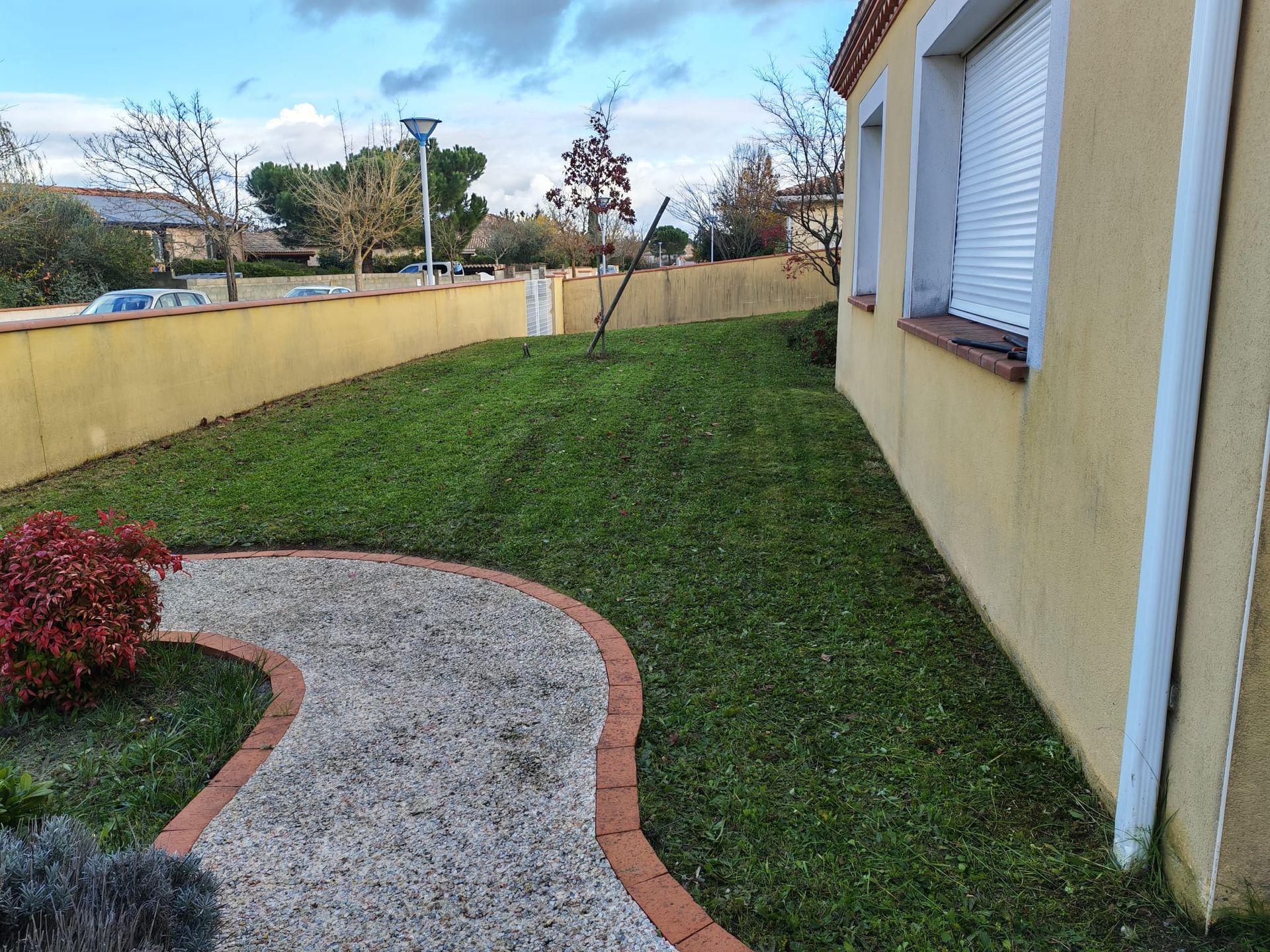 Un chemin de gravier sinueux traverse une petite cour herbeuse jouxtant une maison jaune aux fenêtres à volets blancs.