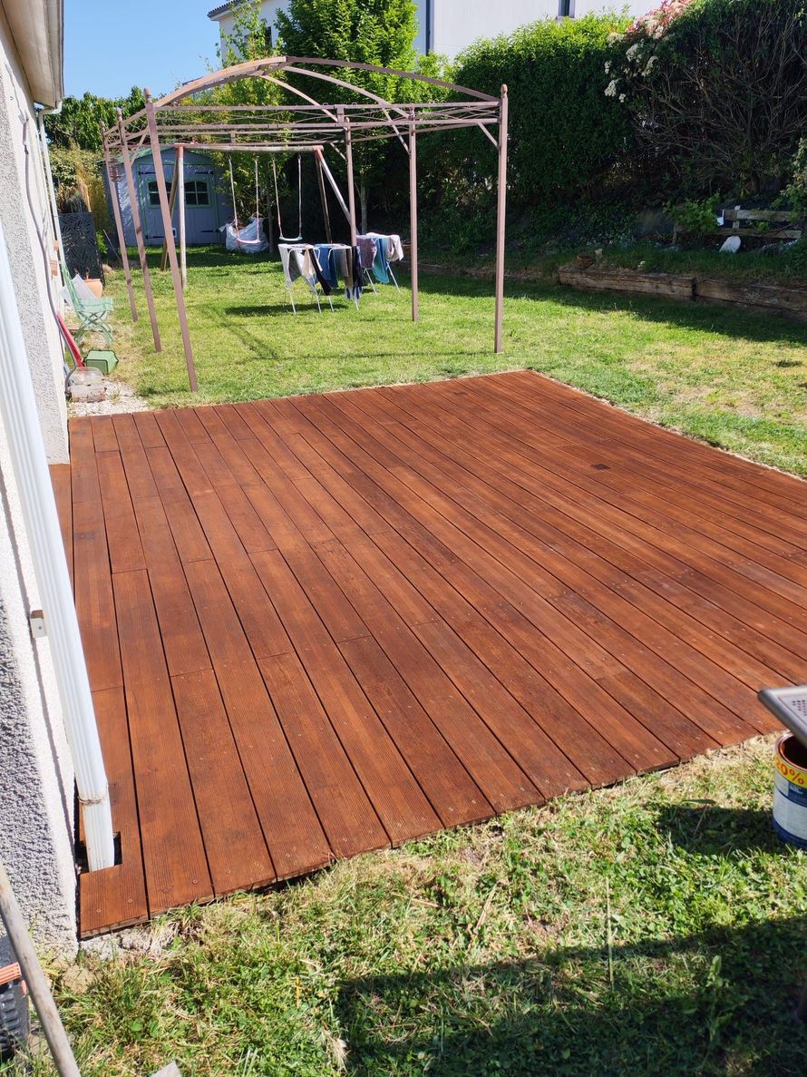 Cour arrière avec une terrasse en bois récemment teintée, jouxtant une pelouse et une structure de pergola.