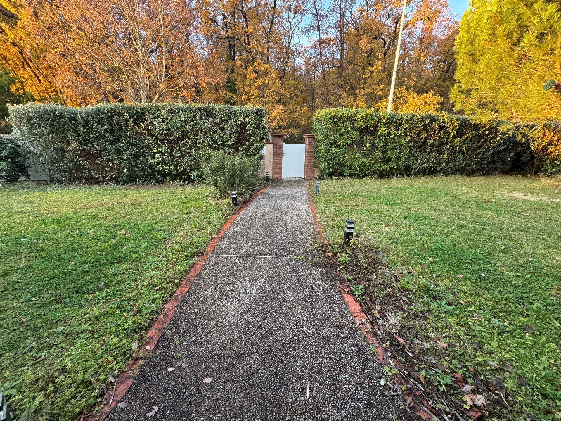 Un étroit chemin de gravier mène à un portail blanc entre des haies, bordé de pelouse verte et d'arbres aux couleurs d'automne.