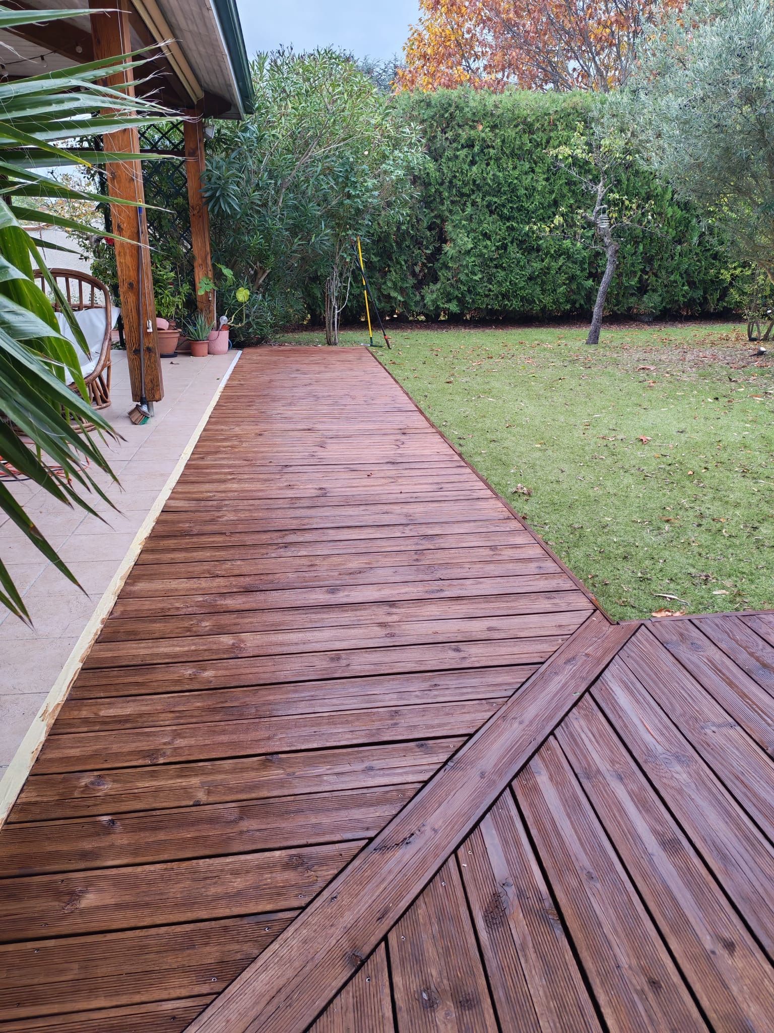 Allée en bois longeant une pelouse verte et une haie, avec des planches trempées par la pluie, et un petit patio.