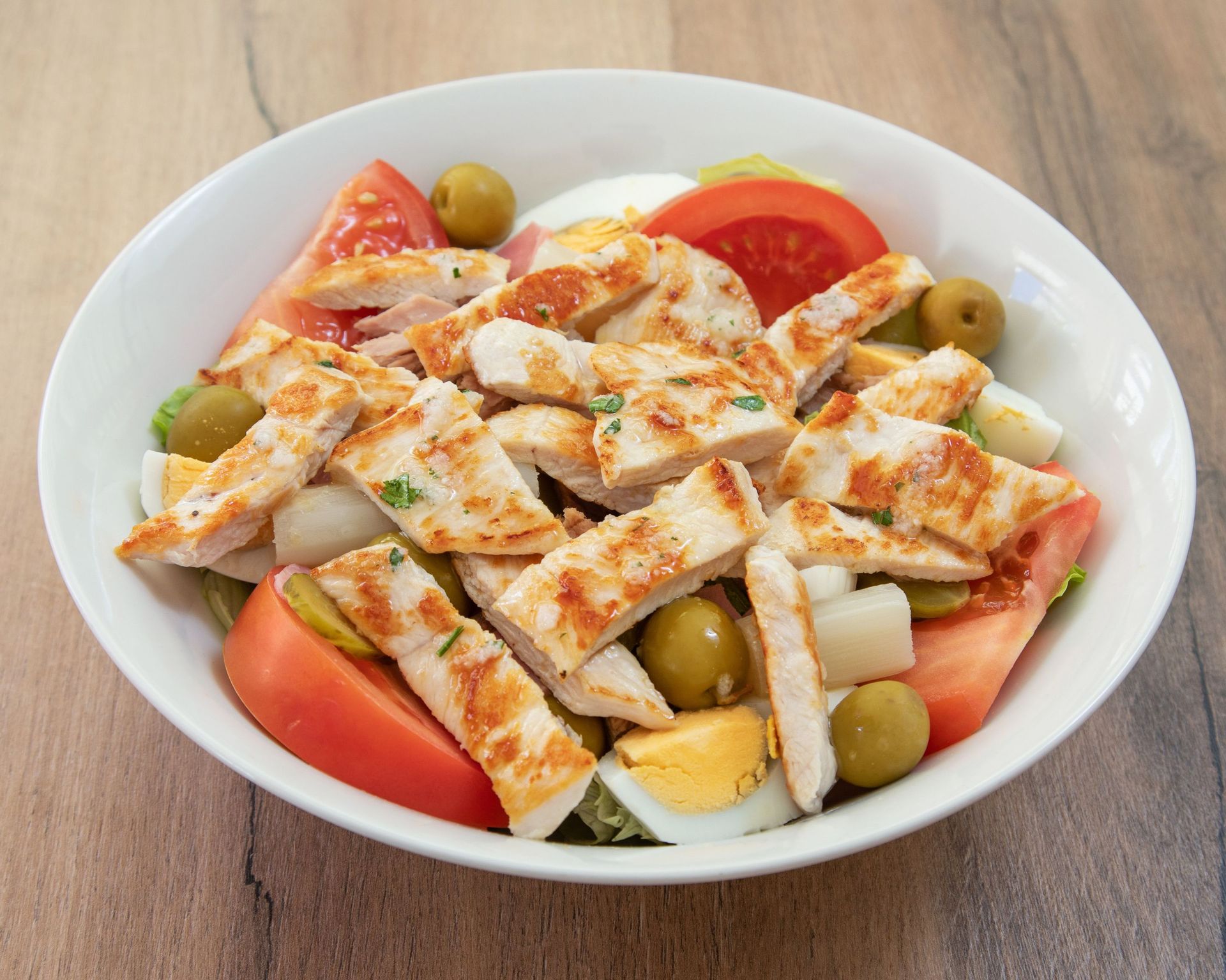 Un tazón blanco lleno de pollo, tomates, aceitunas y huevos.