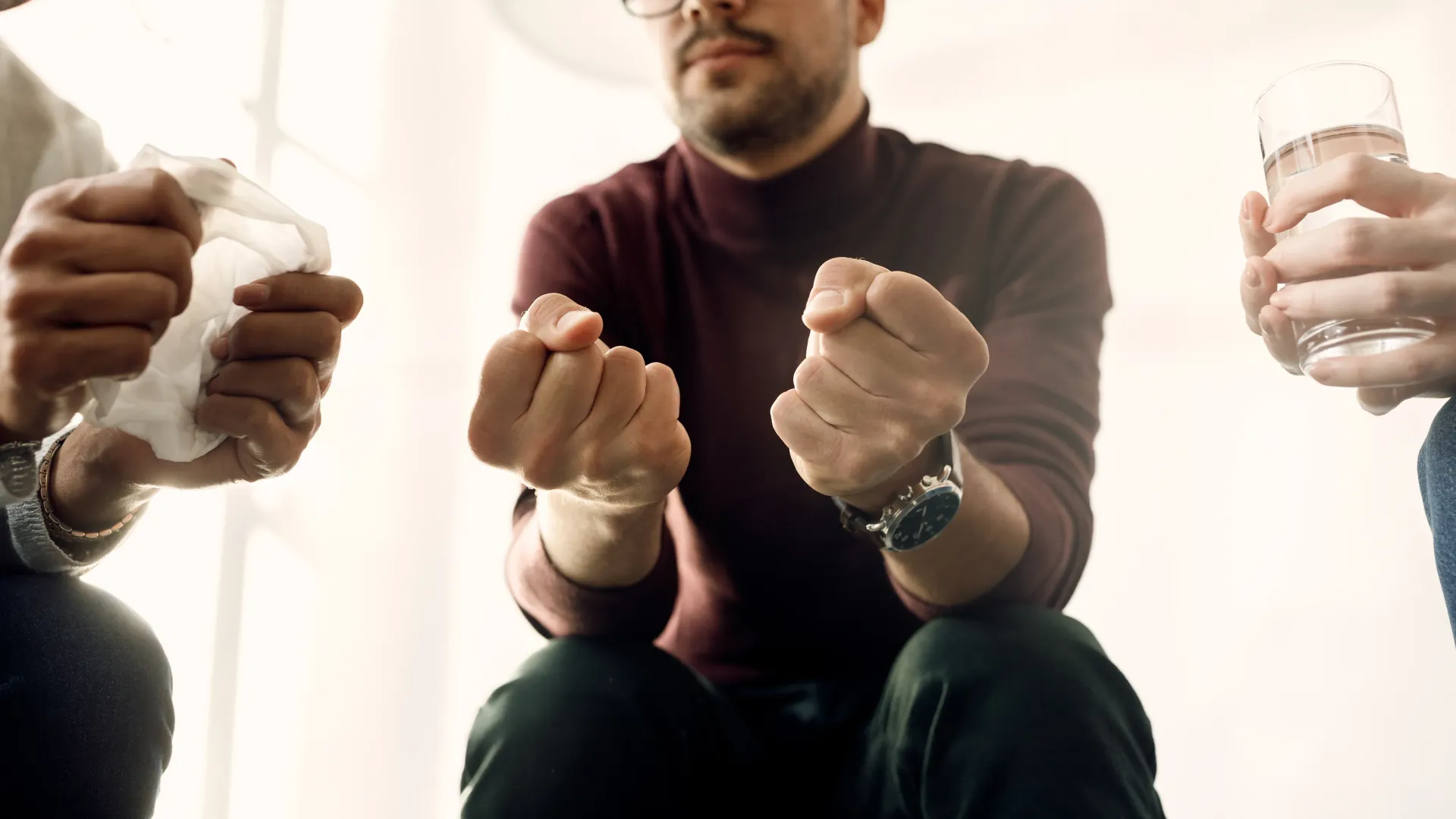 Personas sentadas en círculo, con los puños cerrados y vasos en la mano, en una habitación luminosa.