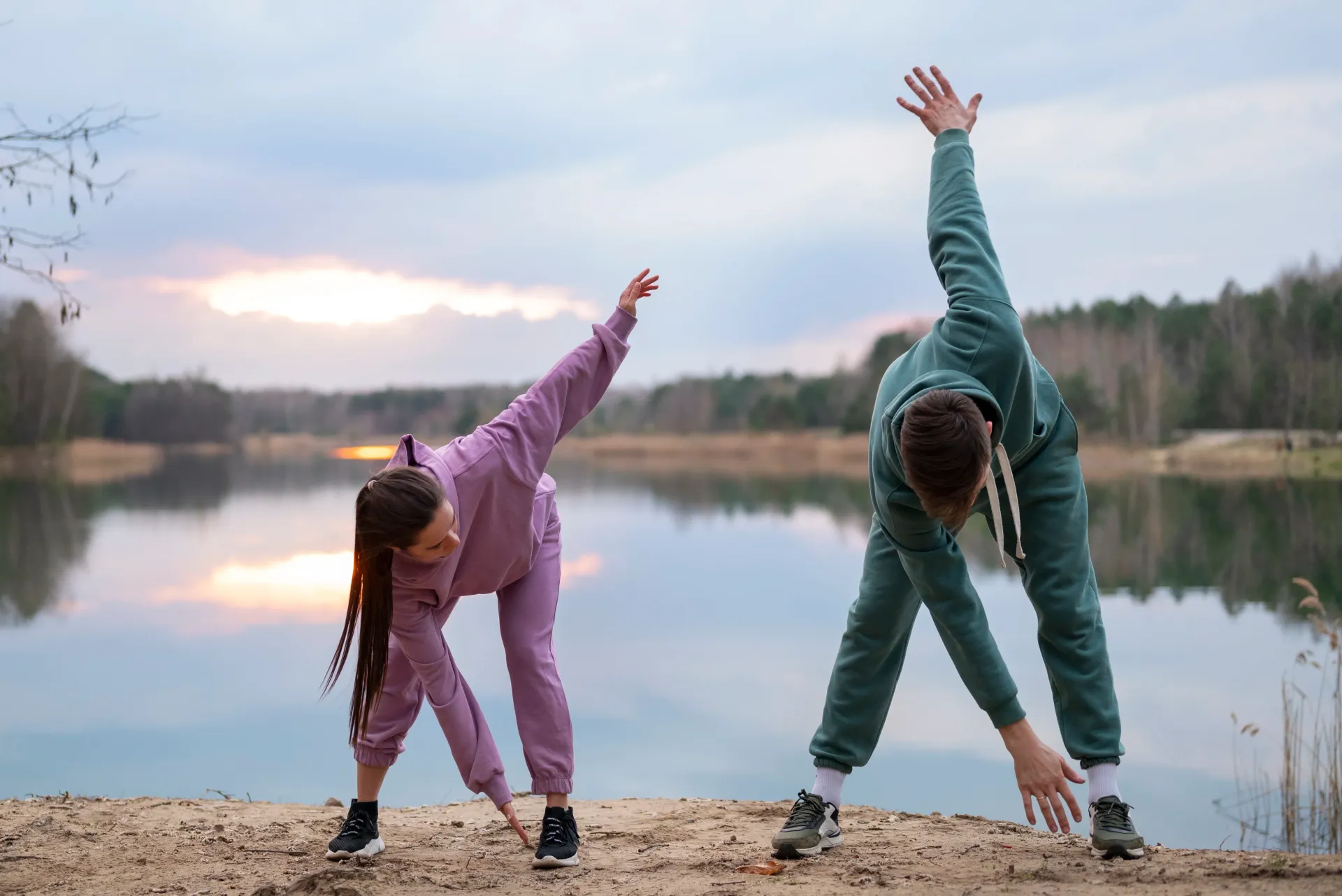 Dos personas se estiran en la orilla de un lago al atardecer, con los brazos extendidos hacia el agua y el cielo.