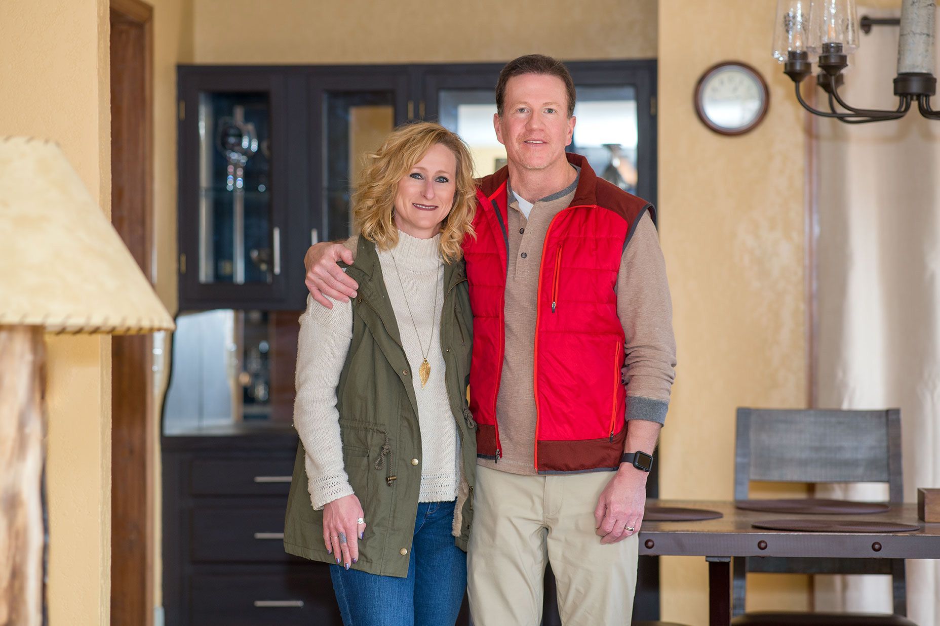 Couple with arms around each other in a room, the man wearing a red vest, the woman wearing a green vest.