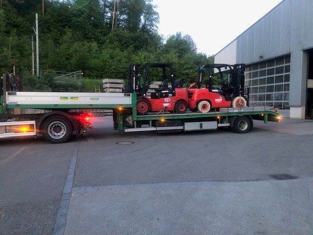 Ein rot-weisser Lastwagen transportiert ein Gerüst und einen Steinblock auf einer Bergstrasse. Bewölkter Himmel.