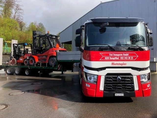 Ein roter und weisser Pickup mit Pritsche, auf der eine Pistenraupe transportiert wird, in einer verschneiten Berglandschaft.