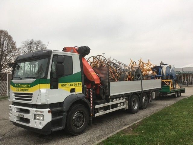 Ein LKW mit orangefarbenem Kran hebt an einer grauen Industriehalle.
