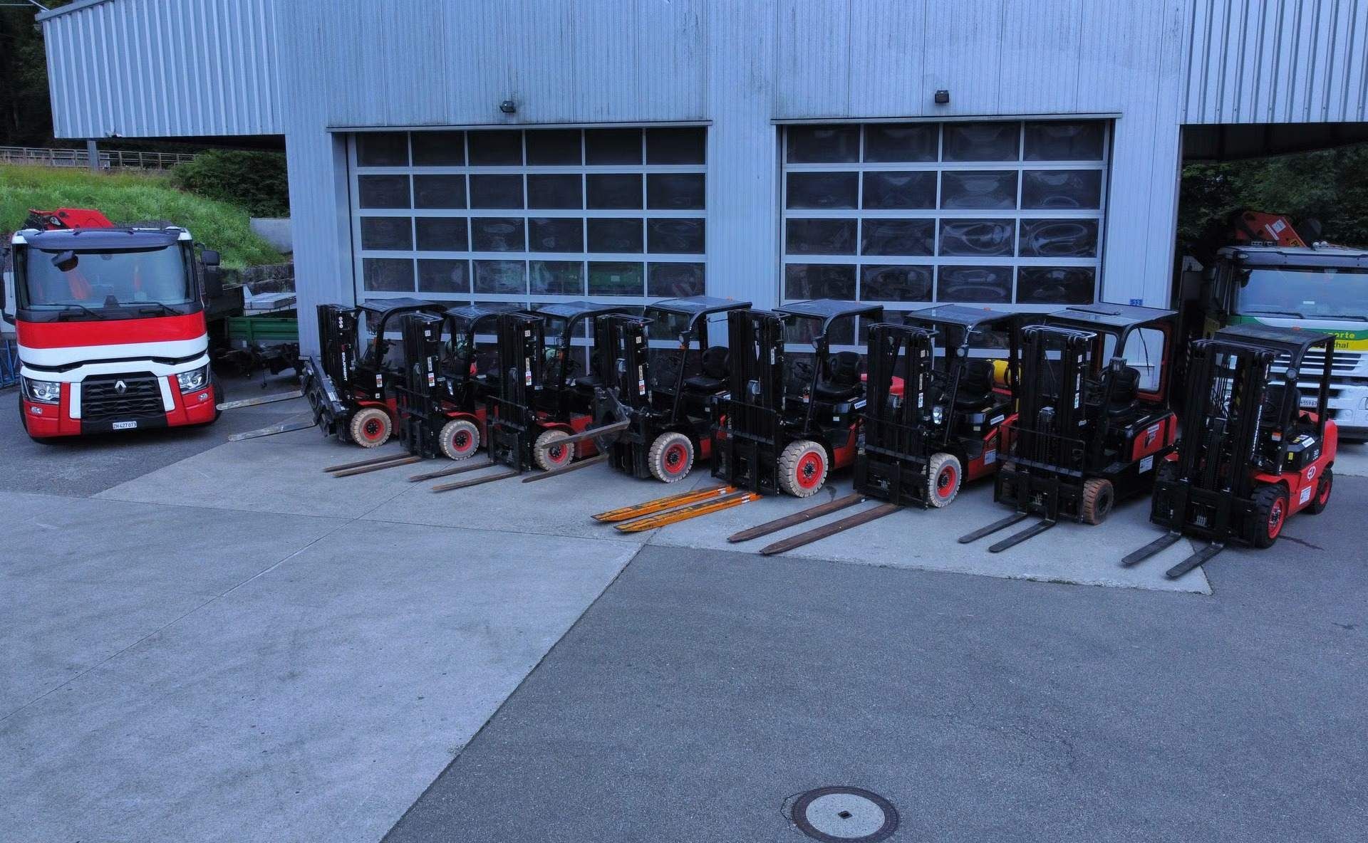 Vor einem Gebäude mit Garagentoren standen mehrere dunkle Gabelstapler in einer Reihe. Links davon befand sich ein rot-weisser Lkw.