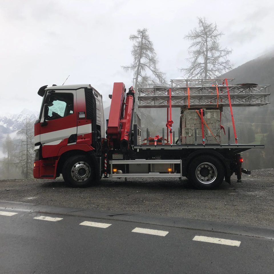 Ein rot-weisser Lastwagen transportiert ein Gerüst und einen Steinblock auf einer Bergstrasse. Bewölkter Himmel.
