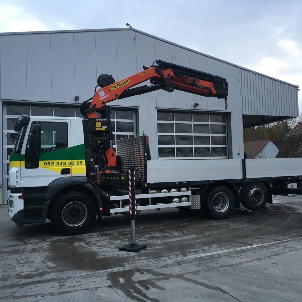 Ein LKW mit orangefarbenem Kran hebt an einer grauen Industriehalle.
