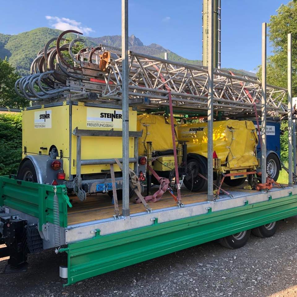 Ein Anhänger, beladen mit gelber und blauer Ausrüstung, einer Metallleiter und einer grünen Ladefläche, vor einer Bergkulisse.