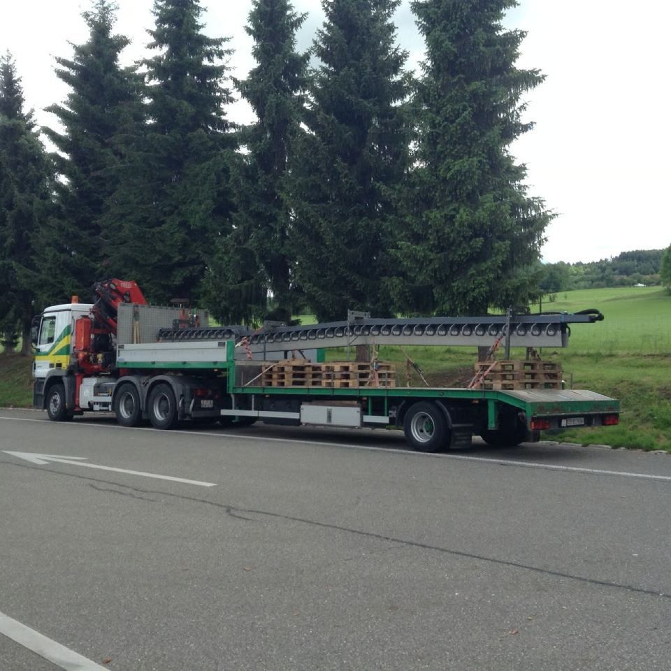 Ein LKW mit Kran, der Baumaterialien auf einem Pritschenanhänger transportiert, steht am Strassenrand.