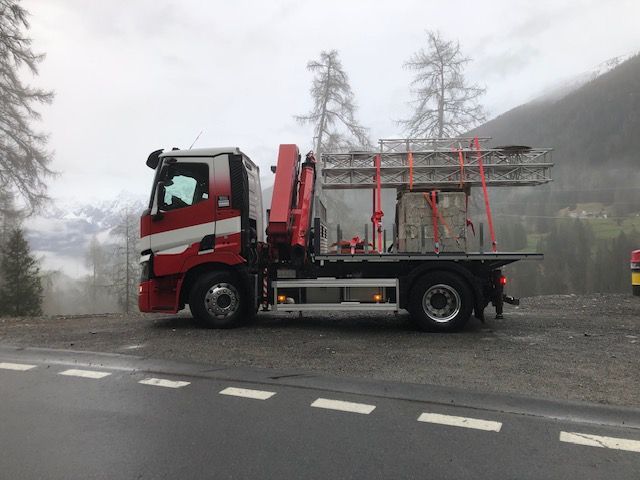 Ein rot-weisser Lastwagen transportiert ein Gerüst und einen Steinblock auf einer Bergstrasse. Bewölkter Himmel.