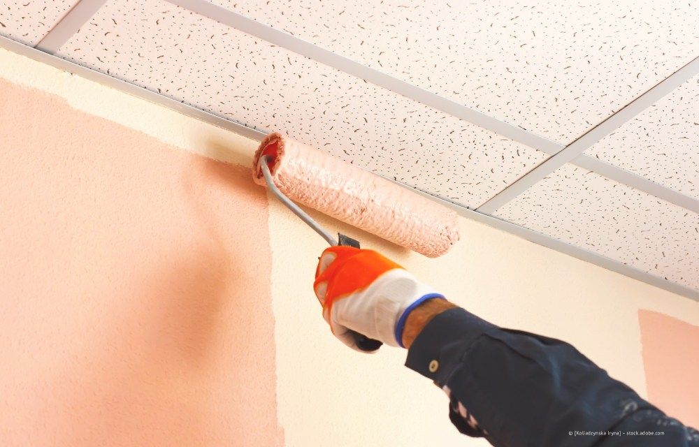 Mit einer Farbrolle rosa Farbe von Hand auf eine Wand in der Nähe einer abgehängten Decke auftragen.