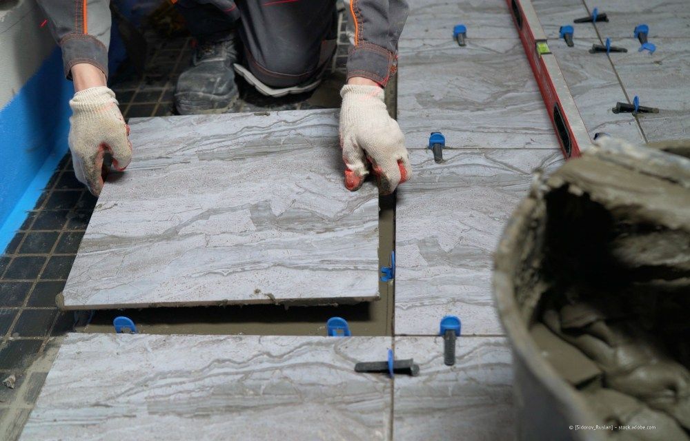 Ein Arbeiter mit Handschuhen verlegt während der Installation eine große graue Fliese auf dem Boden.