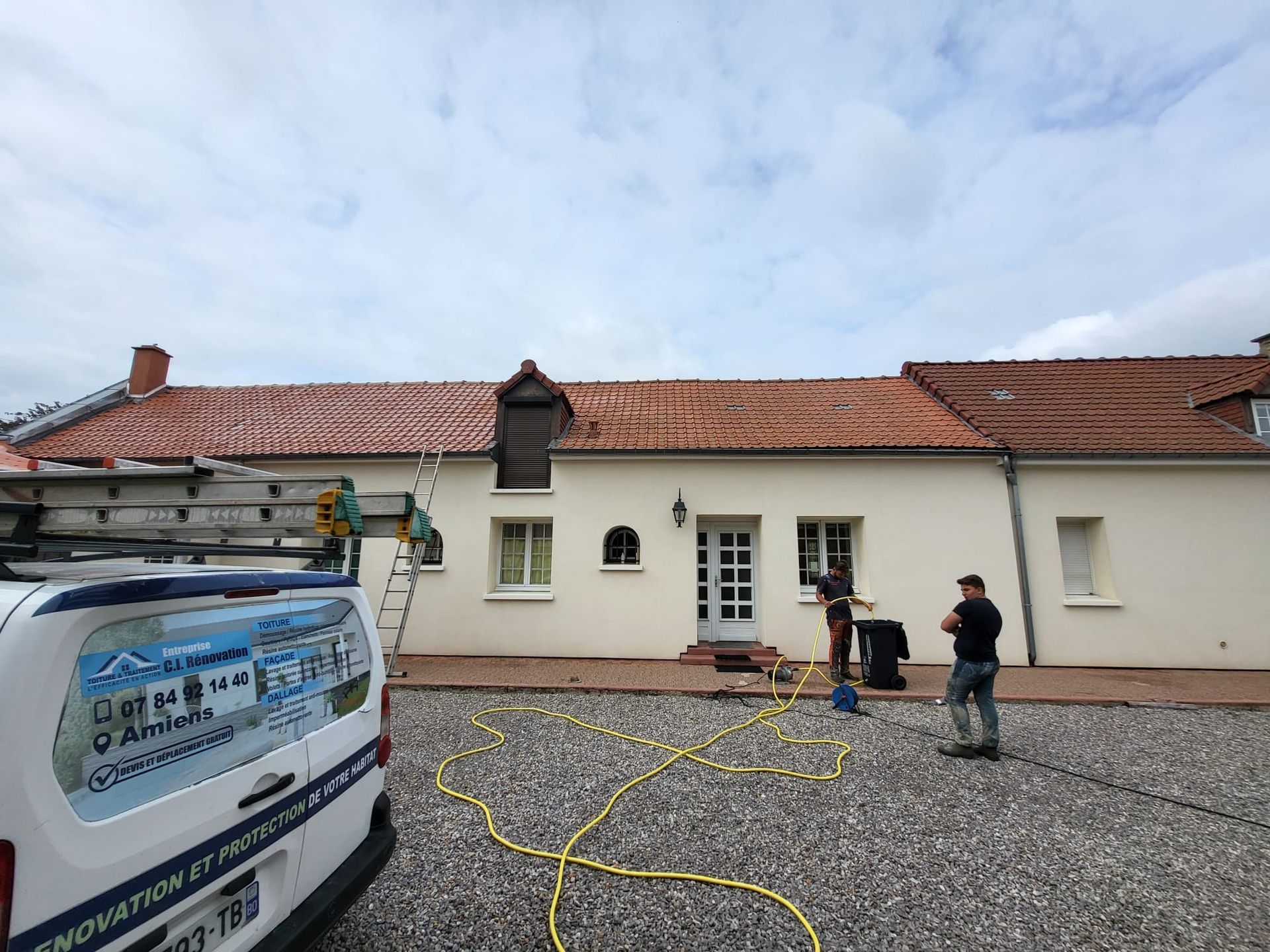 Des professionnels de toiture devant une maison avec une voiture en premier plan et les tuyaux d'arrosage.