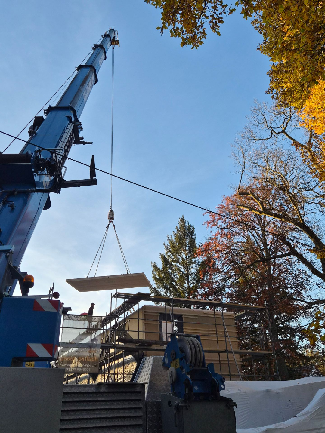 Großer Kran hebt Bauteil zwischen herbstlichen Bäumen in blauem Himmel