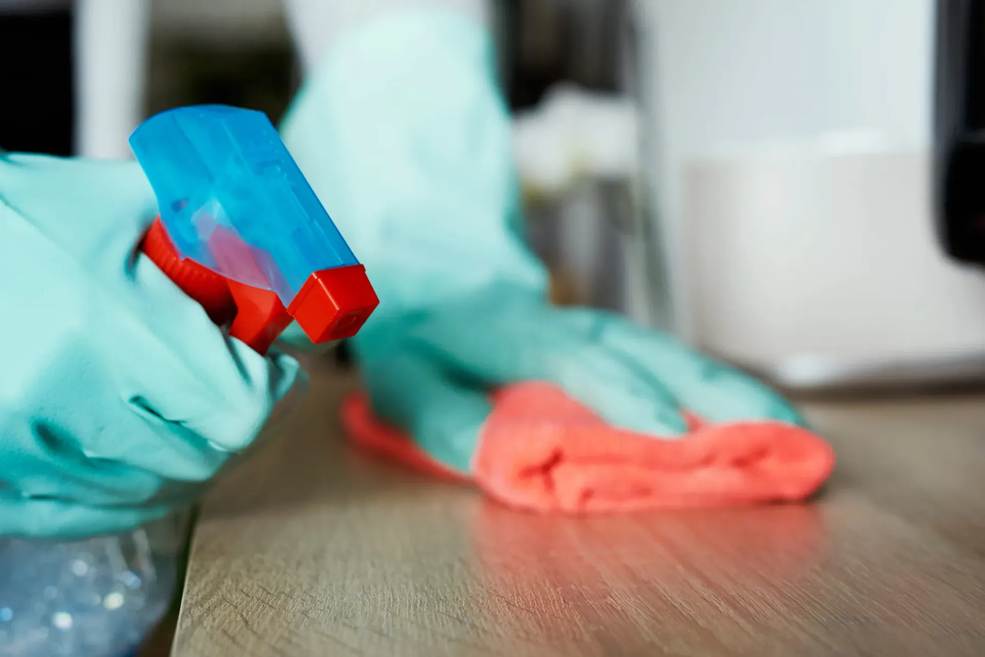 Manos con guantes de goma azul claro limpiando una superficie de madera con un pulverizador y un paño naranja.
