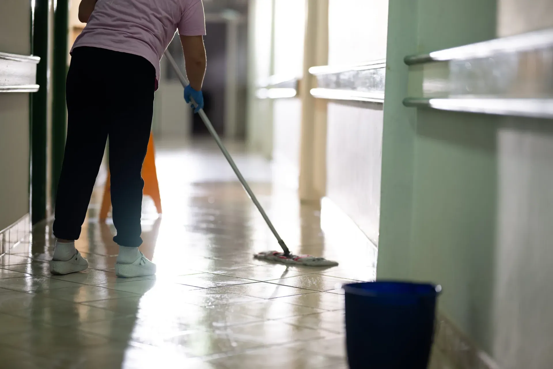 Una persona con una camisa rosa fregando el suelo de baldosas de un pasillo con una fregona mojada y un cubo azul cerca.