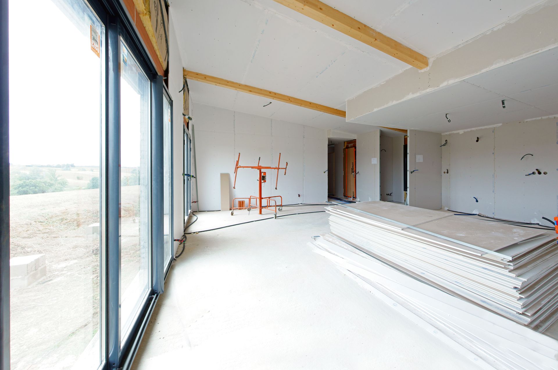 Intérieur d'une pièce en construction, avec de grandes fenêtres, des cloisons sèches et des poutres en bois.