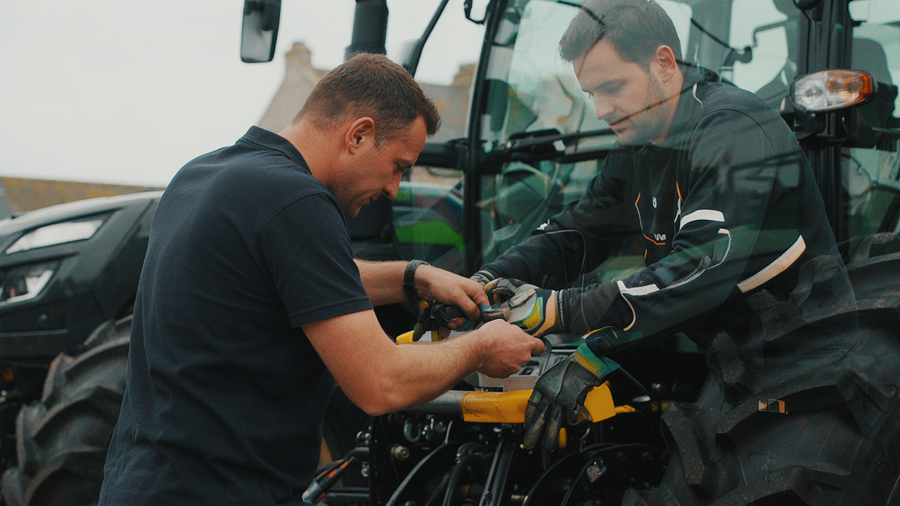 Deux hommes travaillent sur un moteur de moto à l'intérieur d'un véhicule, l'un d'eux se penchant par l'ouverture latérale.